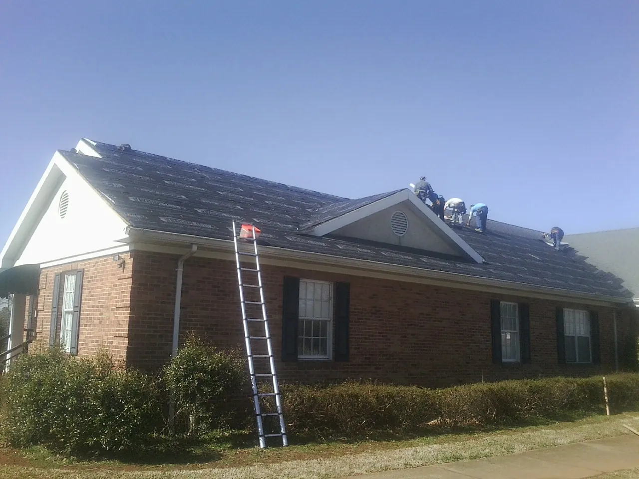 Crew tearing shingles off a brick ranch roof with a ladder set — Emergency Roof Repair work by our crew in East Calais