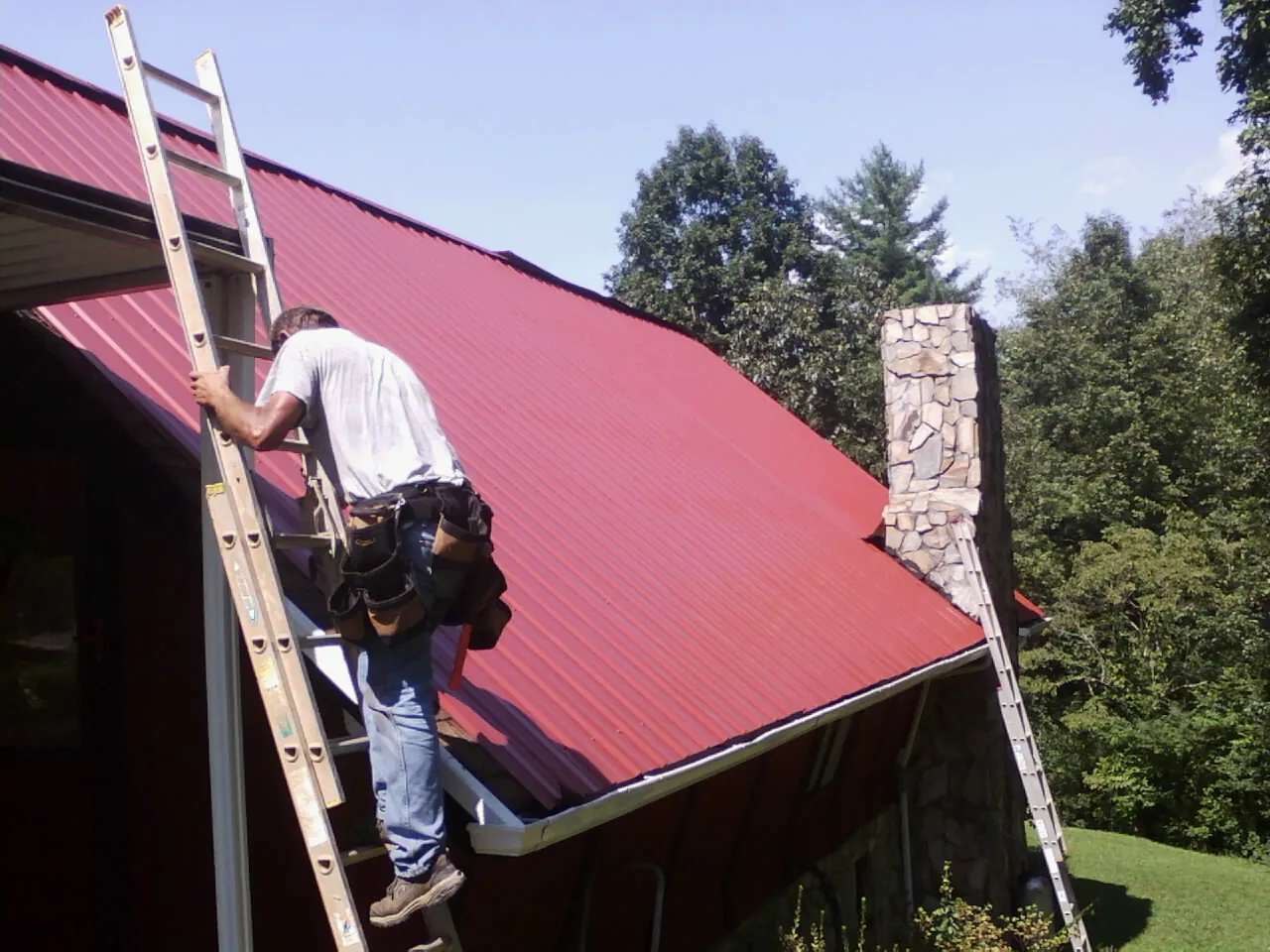 Roofer on a ladder beside a red metal roof and stone chimney — Soffit & Fascia Repair by Roofing Brothers in East Calais